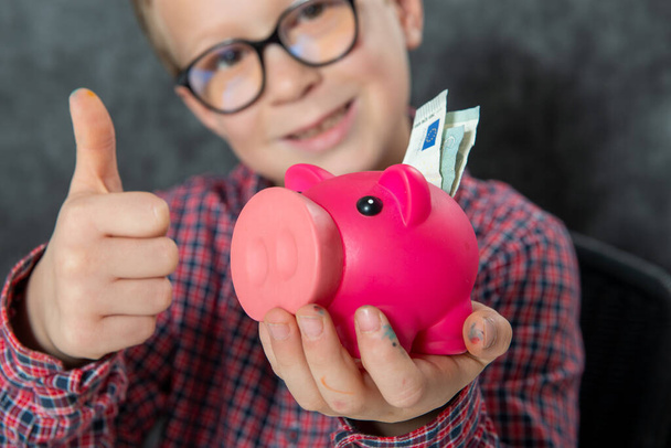 Sorriso menino com óculos coloca dinheiro em um banco porquinho
 - Foto, Imagem