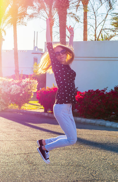 Jovem mulher feliz em óculos de sol está pulando na frente dos arbustos hibiscos vermelhos e rosa florescendo e palmeiras ao pôr do sol
 - Foto, Imagem