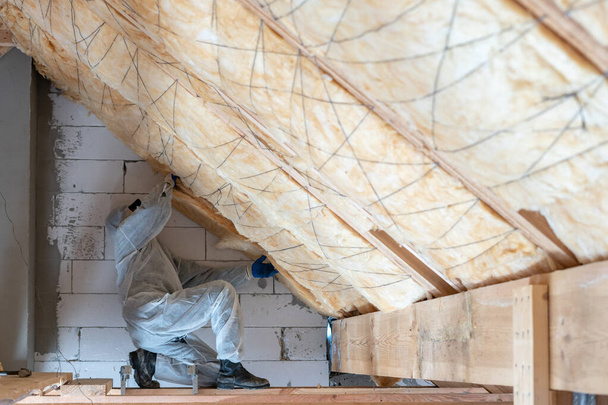 Πλευρική άποψη ενός εργαζομένου με φόρμες εργασίας με πετροβάμβακα μονωτικό υλικό, στέκεται μέσα σε νέο σπίτι υπό κατασκευή στερεώνει στρώμα θερμότητας στην οροφή και τον τοίχο - Φωτογραφία, εικόνα