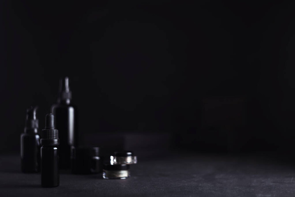 Single sharp small shiny black glass dropper bottle and other blurred skincare makeup packaging on soft matte black background - 写真・画像