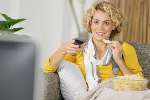 Genç bir kadın televizyon seyrediyor ve kanepede cips yiyor. - Fotoğraf, Görsel