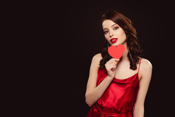 Vonzó fiatal nő piros ruhában tartja szív alakú papír és nézi kamera elszigetelt fekete - Fotó, kép