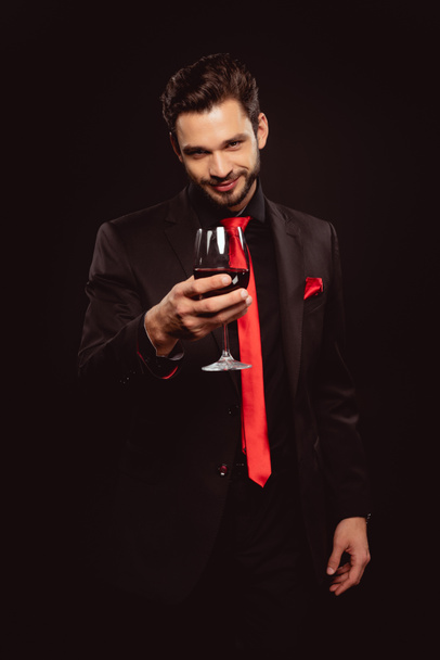 Enfoque selectivo del hombre guapo sosteniendo una copa de vino tinto y mirando a la cámara aislada en negro
 - Foto, Imagen