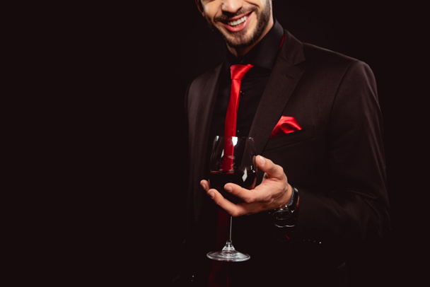 Przycięty widok eleganckiego człowieka uśmiechającego się trzymając kieliszek czerwonego wina na czarno - Zdjęcie, obraz