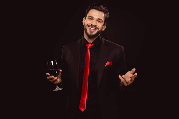 Handsome man in suit and red tie holding glass of wine and smiling at camera isolated on black - Fotoğraf, Görsel