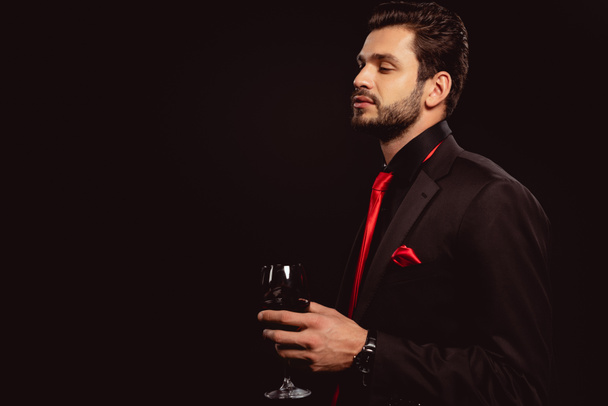 Vista lateral de hombre elegante en traje sosteniendo copa de vino tinto aislado en negro
 - Foto, Imagen