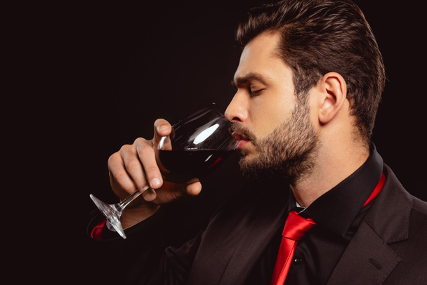 Вид сбоку на красивого мужчину в костюме, пьющего красное вино, изолированное на черном
 - Фото, изображение