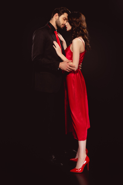 Vista lateral do homem bonito abraçando bela namorada em vestido vermelho isolado no preto
 - Foto, Imagem