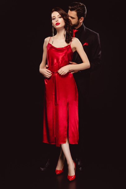 Full length of elegant woman in red dress looking at camera near handsome boyfriend in suit isolated on black - Foto, imagen