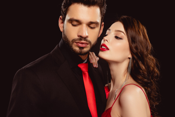 Elegant woman looking at handsome boyfriend in suit and red tie isolated on black - Φωτογραφία, εικόνα