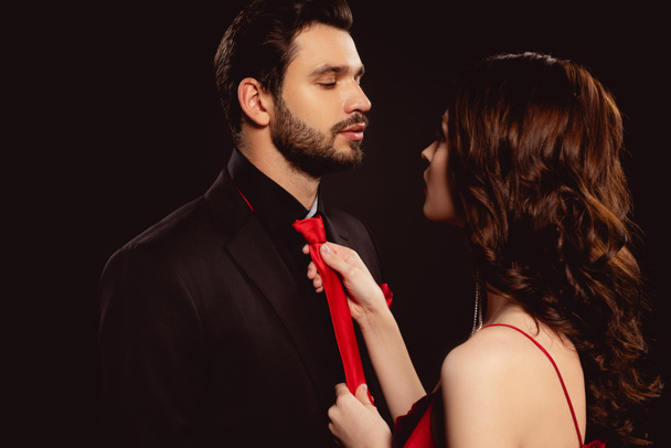Beautiful woman adjusting tie of handsome boyfriend isolated on black - Фото, изображение