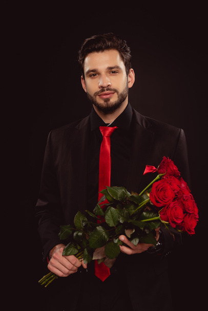 Handsome man in suit holding bouquet of red roses and looking at camera isolated on black - Foto, Imagem