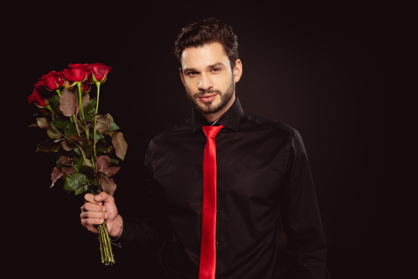 Homem elegante olhando para a câmera e segurando buquê de rosas vermelhas isoladas em preto
 - Foto, Imagem