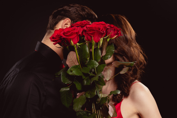Vue latérale du couple élégant couvrant les visages de roses rouges isolées sur noir
 - Photo, image