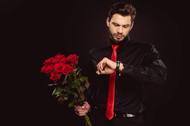 Όμορφος άντρας που κοιτάζει το ρολόι και κρατάει ένα μπουκέτο κόκκινα τριαντάφυλλα απομονωμένα σε μαύρο. - Φωτογραφία, εικόνα