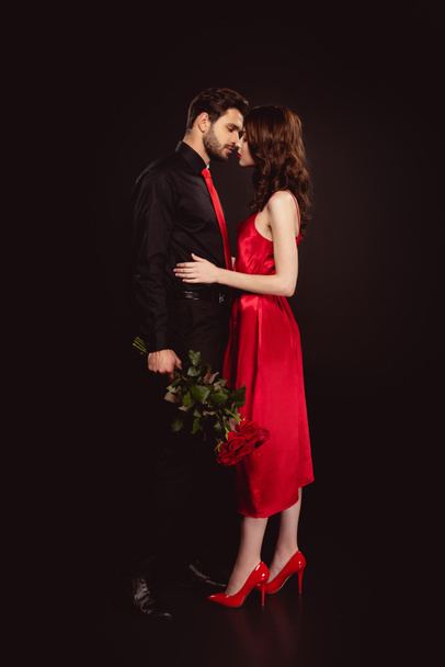 Widok z boku mężczyzna w formalnym stroju gospodarstwa bukiet czerwonych róż i całuje kobieta w sukience izolowane na czarno - Zdjęcie, obraz