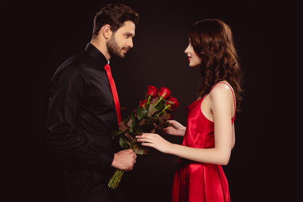 Side view of handsome man giving roses to attractive girlfriend isolated on black - Foto, Imagem
