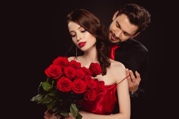 Elegant man taking off dress from girlfriend with red roses isolated on black - Photo, Image