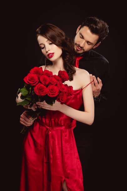 Handsome man taking off dress from elegant girlfriend with red roses isolated on black - Photo, Image