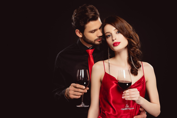 Bir bardak şarapla şık bir kadın, kameraya bakıyor. Yakışıklı erkek arkadaşının yanında, siyahta izole edilmiş. - Fotoğraf, Görsel