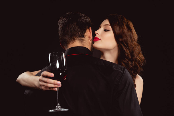 Όμορφη γυναίκα με ένα ποτήρι κρασί που φιλάει το αγόρι της απομονωμένη στα μαύρα. - Φωτογραφία, εικόνα