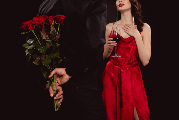 Vista ritagliata di uomo nascondendo mazzo di rose vicino bella fidanzata con vino rosso isolato su nero
 - Foto, immagini