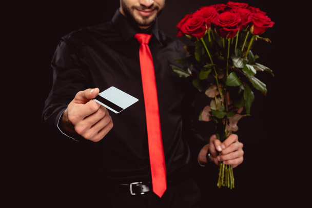 Vista recortada de un hombre elegante con tarjeta de crédito y rosas mientras mira la cámara aislada en negro
 - Foto, Imagen