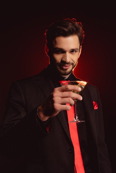 Handsome man in suit holding glass of cocktail on black background with lighting - Fotoğraf, Görsel