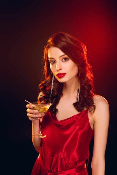 Krásná žena v červených šatech drží sklo koktejlu na černém pozadí s osvětlením - Fotografie, Obrázek