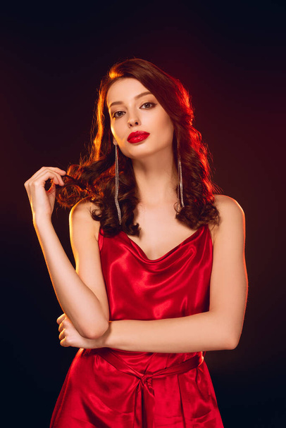 Kırmızı elbiseli çekici bir kız saçlarına dokunuyor ve siyah kameraya bakıyor. - Fotoğraf, Görsel
