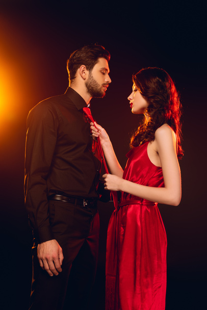 Side view of elegant woman holding tie of boyfriend on black background with lighting - Фото, изображение