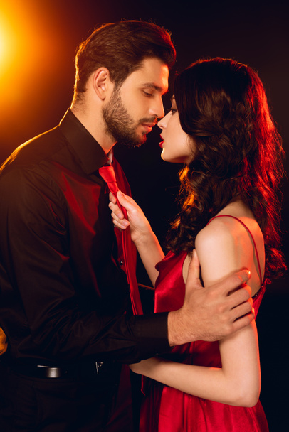 Vista lateral homem elegante beijando bela namorada em vestido vermelho no fundo preto com iluminação
 - Foto, Imagem