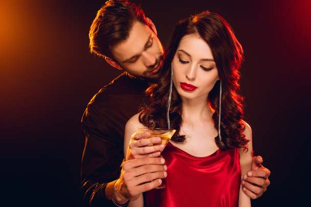 Handsome man embracing elegant girlfriend and holding glass of cocktail on black background with lighting - Photo, Image