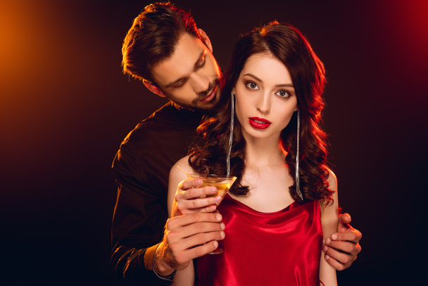 Elegant woman in red dress holding glass of cocktail near handsome boyfriend on black background with lighting - Foto, afbeelding
