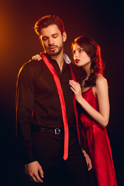 Attractive girl in red dress hugging handsome boyfriend with untied tie on black background with lighting - Photo, Image