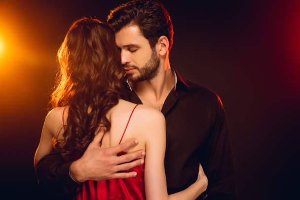 Handsome man embracing girlfriend in red dress on black background with lighting - Foto, imagen