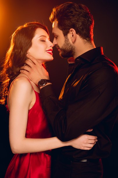 Side view of elegant man touching neck of beautiful smiling girl on black background with lighting - 写真・画像