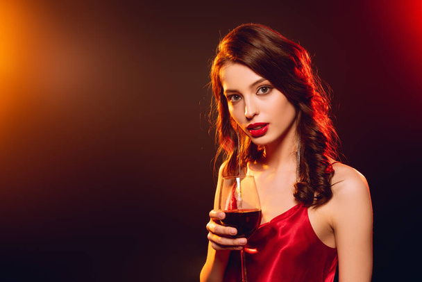 Elegant girl in red dress holding glass of wine and looking at camera on black background with lighting - Photo, Image