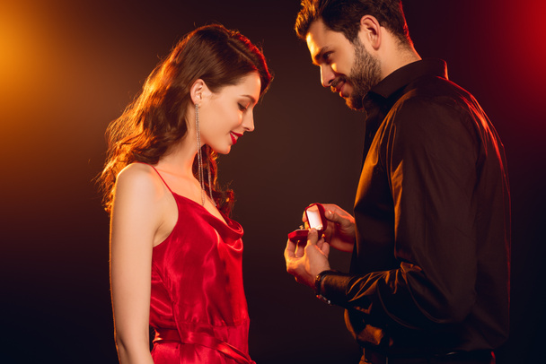 Side view of man holding gift box with jewelry ring near smiling woman in red dress on black background with lighting - Foto, immagini