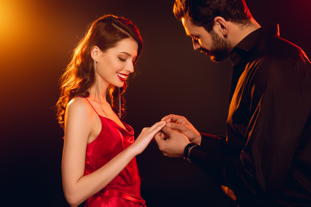 Side view of man putting ring on finger of smiling girlfriend in red dress on black background with lighting - Foto, imagen