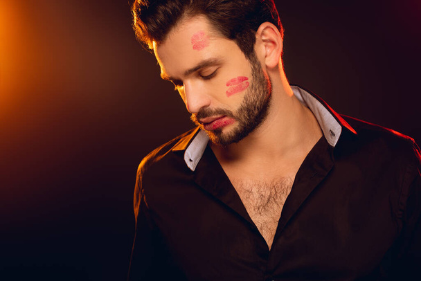 Handsome man with red lipstick prints on face on black background with lighting - Photo, Image