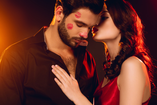 Elegante mujer besando novio guapo con lápiz labial impresiones de la cara sobre fondo negro con iluminación
 - Foto, imagen