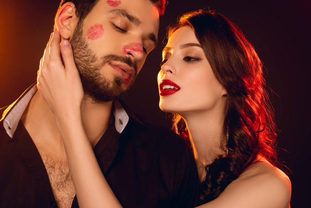 Attractive girl touching handsome boyfriend with lipstick prints on face on black background - Photo, Image