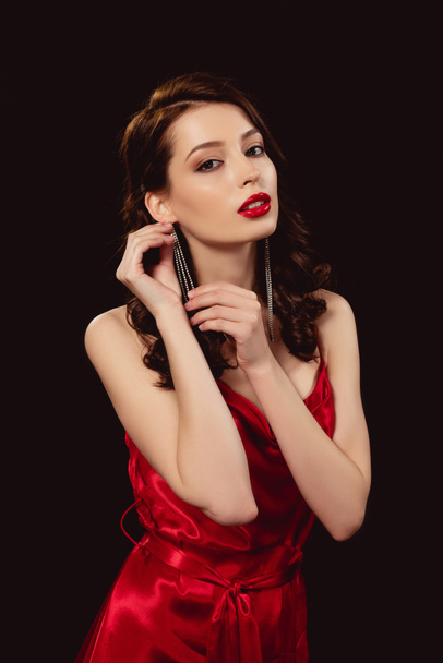 Beautiful woman in red dress touching earring and looking at camera isolated on black - Фото, изображение