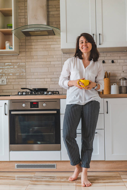 Młody ładny stylowy kobieta trzyma żółty kubek w białej kuchni. gospodyni domowa koncepcja - Zdjęcie, obraz