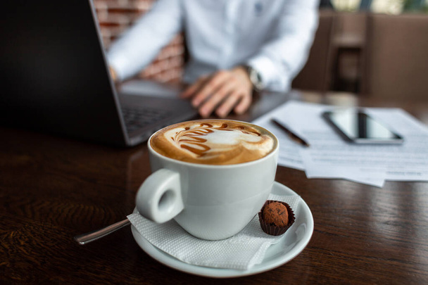 White cup of coffee and a sweet on a saucer on the table at a cafe. Young businessman with a laptop on the background. Freelance and selfeployement concept - Фото, изображение