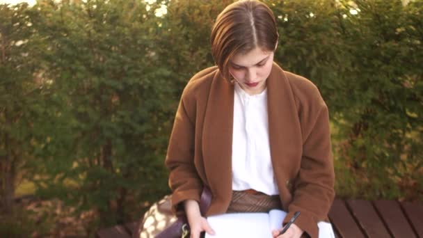 Young girl student preparing for exams in a park on a bench. Brown coat, short haircut. Notebooks and books - Filmagem, Vídeo