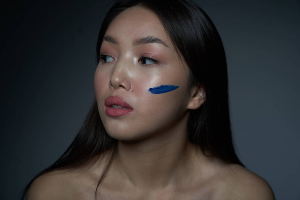  Profilo di una giovane bella ragazza asiatica con una guancia imbrattata di vernice ad olio blu sotto forma di una striscia orizzontale. Foto di primo piano. Su uno sfondo grigio, un luogo da copiare
                               - Foto, immagini