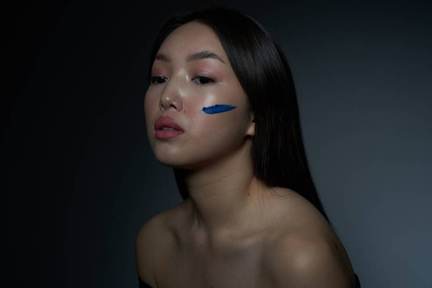  Profilja egy fiatal gyönyörű ázsiai lány egy arc bekent kék olajfesték formájában egy vízszintes csík. Egy közeli kép. A szürke háttér, a hely, hogy másolja                               - Fotó, kép