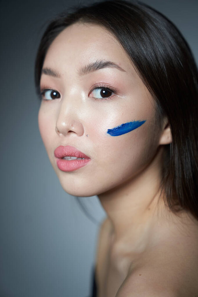  Profile of a young beautiful Asian girl with a cheek smeared with blue oil paint in the form of a horizontal stripe. Close-up photo. On a gray background, a place to copy                               - Fotó, kép
